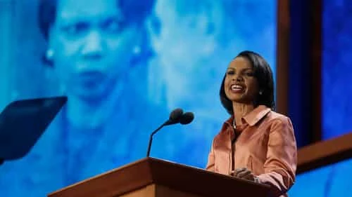 Condoleezza Rice standing behind a wooden podium on an indoor stage, addressing an audience. Condoleezza Rice standing behind a wooden podium on an indoor stage, addressing an audience.