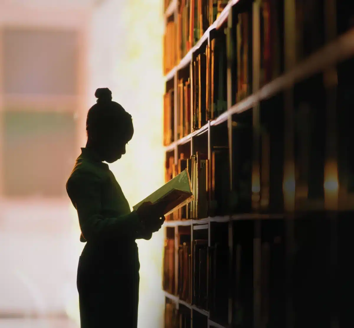 A silhouette of a young reader standing indoors beside a tall wall of bookshelves filled with neatly arranged books. A silhouette of a young reader standing indoors beside a tall wall of bookshelves filled with neatly arranged books.