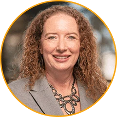 A photo of Tina Burke. The image shows a professional head‑and‑shoulders portrait presented inside a circular frame with a thin orange border, set on a transparent checkerboard background. A single adult is centered and faces forward, wearing a light‑colored blazer and a statement necklace with interlinked shapes. The background inside the circle is softly blurred in warm, neutral tones, suggesting an indoor setting with even, natural lighting that creates a polished, approachable appearance.