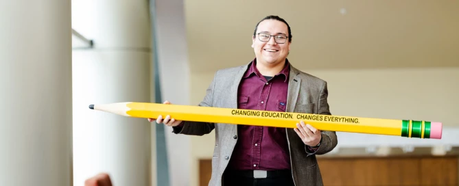 Attendee standing indoors holding an oversized yellow pencil printed with “Changing education changes everything,” in a bright lobby or event space