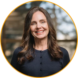 A photo of Denise Inman. The image shows a professional head‑and‑shoulders portrait inside a circular frame with an orange border on a transparent background. A single person faces forward wearing a dark, long‑sleeved top with front buttons. Hair falls past the shoulders, and the background inside the circle is softly blurred with warm, neutral tones.